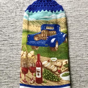 May include: A blue and white kitchen towel with a crocheted blue top. The towel features a vintage blue pickup truck with barrels in the back, a picnic scene with wine glasses, grapes, and cheese, and the text "Wine Country".