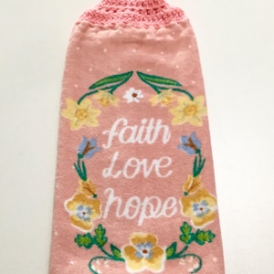 May include: Pink kitchen towel with a crocheted top and a floral design. The towel has the words "faith love hope" in a wreath of flowers.