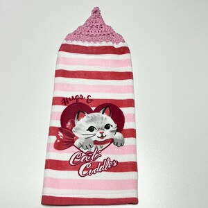May include: Pink and white striped kitchen towel with a crocheted pink top. The towel features a white cat with a red bow in a red heart with the text "Hugs & Cat Cuddles".