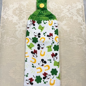 May include: A white kitchen towel with a green crocheted hanging loop and a yellow button. The towel features a repeating pattern of Mickey Mouse and Minnie Mouse faces, shamrocks, horseshoes, and green hats.