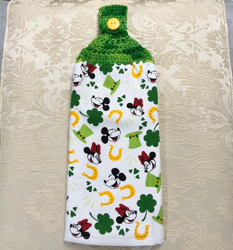 May include: A white kitchen towel with a green crocheted hanging loop and a yellow button. The towel features a repeating pattern of Mickey Mouse and Minnie Mouse faces, shamrocks, horseshoes, and green hats.