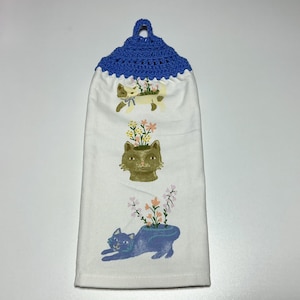 May include: A white kitchen towel with a blue crocheted hanging loop. The towel features a design of three cats with flowers growing out of their heads.