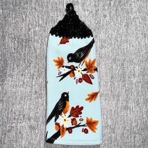 May include: A light blue kitchen towel with a black, glittery top. The towel features two black birds with orange chests perched on branches with orange and brown leaves and white flowers.