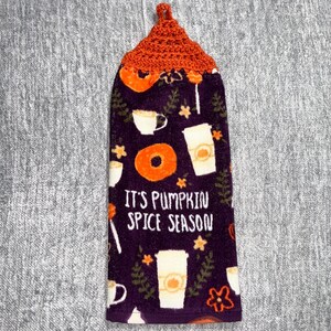 May include: A decorative kitchen towel with a dark purple background and an orange crocheted top. The towel features a pattern of coffee cups, donuts, and the text "IT'S PUMPKIN SPICE SEASON".