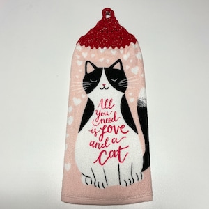 May include: Pink kitchen towel with a black and white cat illustration. The cat is sitting and has the text "All you need is love and a cat" printed on it. The towel has a red crocheted hanging loop.