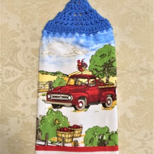 May include: A hanging kitchen towel featuring a blue crocheted top with a loop. The fabric displays a red vintage pickup truck on a farm with a rooster, green trees, and baskets of red apples. A thin red horizontal stripe accents the bottom edge.