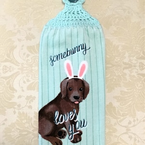 May include: A light blue kitchen towel with a brown dog wearing bunny ears. The dog is embroidered with the text "somebunny loves you". The towel has a crocheted loop with a button at the top.