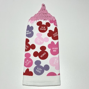 May include: A white kitchen towel with a pink crocheted hanging loop. The towel features a repeating pattern of red, pink, and purple Mickey Mouse ears with Valentine's Day themed messages like "Be Mine", "UR Cute", "Cutie Pie", "Luv You", "XOXO", and "Kiss Me".
