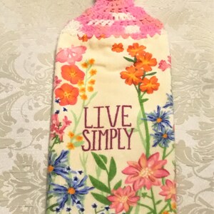 May include: A white kitchen towel with a floral print and the text "Live Simply". The towel has a pink crocheted loop with a button at the top.
