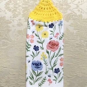 May include: A white hanging kitchen towel featuring a colorful floral print with pink roses, blue flowers, yellow daisies, and small bees. The top of the towel has a bright yellow crocheted topper with a loop for hanging on a cabinet or oven handle.