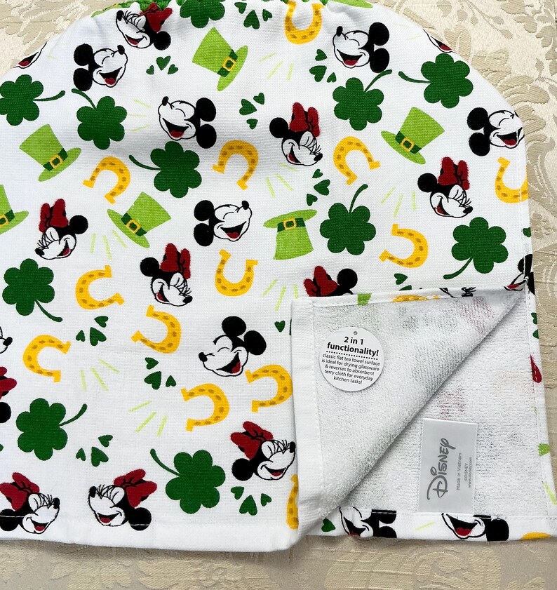May include: White kitchen towel with a green shamrock, gold horseshoe, and Mickey Mouse and Minnie Mouse print. The towel has a white terry cloth side and a white cotton side with the print. The towel has a tag that says "2 in 1 functionality! Cotton towel for drying dishes and a terry cloth towel for everyday kitchen tasks."