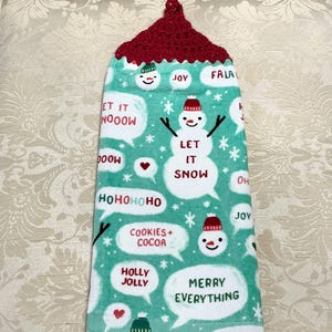 May include: A green kitchen towel with a red crocheted hanging loop. The towel features a snowman design with the text "Let it snow" and other Christmas-themed phrases like "Ho ho ho" and "Merry Everything".