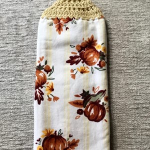 May include: A white kitchen towel with a yellow and white striped pattern and a fall-themed print of pumpkins, leaves, and flowers. The towel has a crocheted beige hanging loop.
