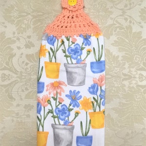 May include: A peach-colored crocheted dish towel holder with a yellow button. The towel has a floral print with blue, yellow, and pink flowers in pots.