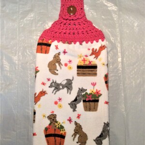 May include: A pink crocheted dish towel holder with a brown button. The towel features a white background with a repeating pattern of dogs and flowers in a variety of colors.