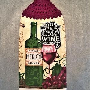 May include: A white kitchen towel with a crocheted burgundy hanging loop. The towel features a vintage-style illustration of a bottle of red wine, a glass of red wine, and grapes. The text on the towel reads "Vintage Sweet Red Wine" and "Vintage Merlot Red Wine".