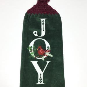 May include: Green kitchen towel with the word "JOY" in white letters with a cardinal bird in the "O". The towel has a burgundy crocheted loop with a white button at the top. The bottom of the towel says "to the world".