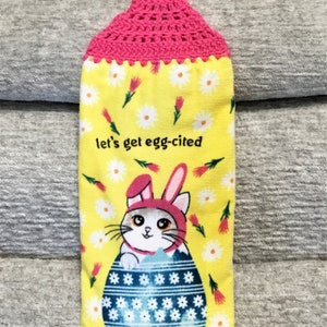 May include: A yellow kitchen towel with a pink crocheted hanging loop. The towel features a white cat wearing a pink bunny ear headband, peeking out of a blue and white patterned egg. The text "let's get egg-cited" is printed on the towel.