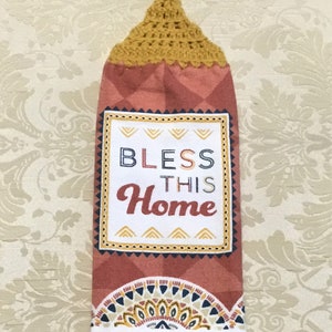 May include: A kitchen towel with a crocheted hanging loop. The towel is red with a white border and features a yellow, black, and white geometric design. The text "Bless This Home" is printed in the center of the towel.