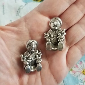 May include: Two silver figurines of people sitting with their arms outstretched, holding smaller figurines of people. The figurines have detailed facial features and are made in a folk art style.