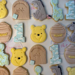 May include: A selection of Winnie the Pooh themed biscuits. Featuring Pooh Bear, Eeyore, honey pots, and the number one. Some biscuits have the name "Arlo" and "Happy Birthday" written on them. The biscuits are decorated with yellow, blue, and brown icing.