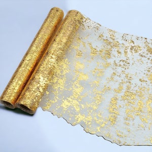 May include: Two rolls of gold-colored decorative fabric. The fabric has a textured, metallic appearance with a distressed design. The rolls are partially unrolled, revealing the fabric's pattern and texture. The fabric is likely used for crafts or event decor.