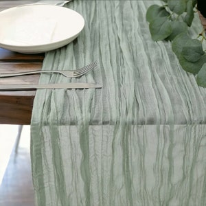 May include: A sage green table runner with a crinkled texture. The runner is laid on a wooden table with a white plate and silverware.