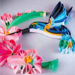 May include: A colorful paper craft of a hummingbird with blue, green, and yellow accents, perched on a pink flower with green leaves.