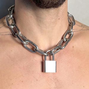 May include: A silver chain necklace featuring large oval links and a square padlock clasp. The necklace is worn around the neck. A striking piece of jewellery.