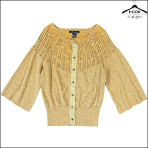 Puede incluir: Rebeca dorada con cuello redondo y mangas tres cuartos. El suéter presenta un canesú con lentejuelas y una botonadura frontal. El logotipo de Hook & Hanger está en la esquina superior derecha. La rebeca es una talla S.