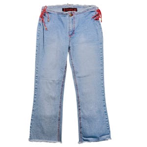 Op de afbeelding: Lichtblauwe denim jeans met rode stiksels en rafelige zomen. De jeans hebben rode veters aan de zijkanten en een knoopsluiting. De tekst "Angel" is zichtbaar op de tailleband.