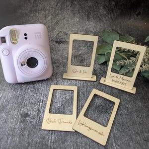 Könnte beinhalten: Eine hellviolette Instax i12 Kamera liegt neben vier Holz-Fotorahmen-Ausschnitten. Die Rahmen sind mit Text graviert, darunter "Du & Ich", "Lisa & Marian 15.09.2025", "Beste Freunde" und "Lieblingsmensch".