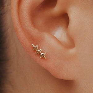 Pode incluir: Um brinco dourado com três elementos conectados em forma de estrela dispostos numa fila linear. A peça de joalharia está posicionada no lóbulo da orelha, exibindo um acabamento metálico polido e um design de estrela com várias pontas.