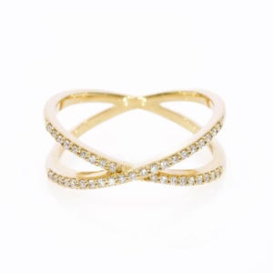 Puede incluir: Un anillo de oro con un diseño cruzado en forma de X. Las dos bandas se cruzan en el centro y están incrustadas con pequeñas piedras claras de corte redondo a lo largo de los bordes exteriores del marco de metal dorado, creando un brillo.