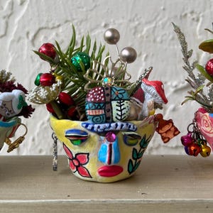 May include: Three whimsical, hand-painted ceramic face planters, each adorned with festive holiday decorations. The planters feature colorful faces, and are filled with miniature ornaments, greenery, and bows, creating a unique holiday display.