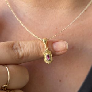 May include: A gold chain necklace featuring a small, elongated hexagonal pendant with a sunburst-style engraved texture and a single, rectangular, deep pink gemstone set vertically in the center of the pendant.