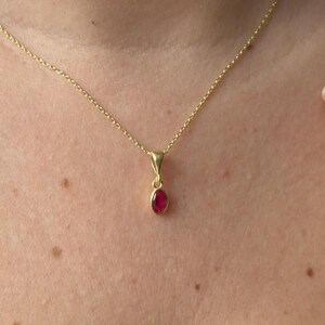 Könnte beinhalten: Eine zarte Goldkettenhalskette mit einem kleinen, ovalen Anhänger, der einen leuchtend roten Edelstein in einer glatten Goldfassung enthält. Die Halskette liegt auf dem Schlüsselbein und zeigt ein schlichtes Design mit einer dünnen, polierten Gliederkette.