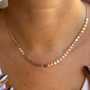 May include: A delicate rose gold necklace with a series of small, round, flat charms. The necklace is worn around a person's neck, highlighting its simple yet elegant design. The chain and charms have a polished finish, reflecting light.