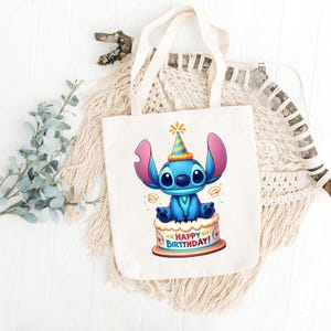 May include: A cream-colored tote bag featuring a cartoon illustration of Stitch from Lilo & Stitch. Stitch is blue with large ears and is sitting on a birthday cake. The cake has the words "HAPPY BIRTHDAY!" on it. The bag has cream-colored handles.