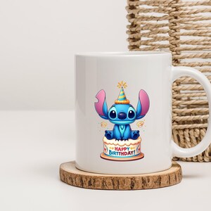 May include: White ceramic mug featuring a blue cartoon character with large ears and a party hat, sitting atop a birthday cake. The cake has the words "HAPPY BIRTHDAY!" on it. The mug sits on a wooden coaster.