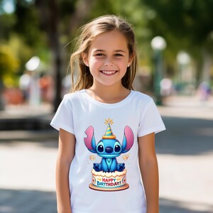 May include: White t-shirt featuring a cartoon character, Stitch, sitting on a birthday cake. The character is blue with large ears and wearing a party hat. The cake has the words "Happy Birthday!" on it.