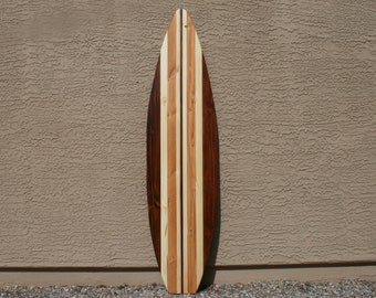 May include: A wooden surfboard with a brown, white, and natural wood grain pattern. The surfboard is leaning against a textured wall.