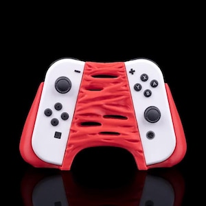 Puede incluir: Un accesorio de mando de videojuegos rojo y blanco. El accesorio tiene una sección central roja texturizada con múltiples aberturas y secciones de mando blancas a ambos lados. El mando tiene botones negros y almohadillas direccionales.