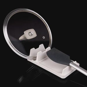 May include: A white rectangular kitchen utensil rest with multiple slots, holding a circular glass pot lid with a metal rim and a gray silicone spatula with a silver handle. The organizer is designed to keep countertops clean while cooking.