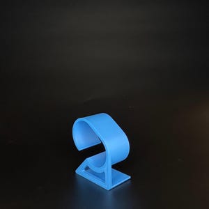 Puede incluir: Un soporte de plástico azul claro para un reloj inteligente. El soporte tiene forma de C con una base plana.