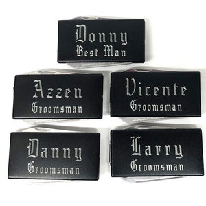 May include: Four black pocket knives with silver engraved lettering. The knives are engraved with the names "Donny Best Man", "Azzen Groomsman", "Vicente Groomsman", and "Danny Groomsman".