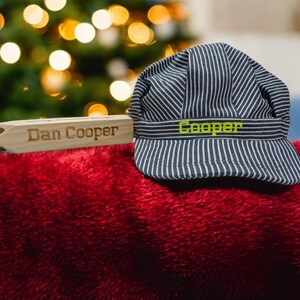 May include: A wooden whistle with the text "Dan Cooper" engraved on it, next to a blue and white striped engineer's cap with the word "Cooper" embroidered in green on the front.