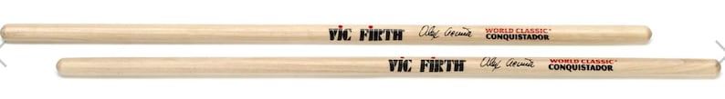 Puede incluir: Dos baquetas de bater&iacute;a de madera clara, con la inscripci&oacute;n "VIC FIRTH" y "WORLD CLASSIC CONQUISTADOR". Las baquetas son de color marr&oacute;n claro y tienen una firma.