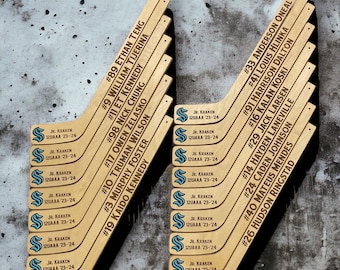 May include: A set of wooden hockey sticks with player names and numbers printed on them. The sticks are arranged in two columns, with the names and numbers facing outward. The sticks are all the same size and shape, and they are made of a light brown wood.