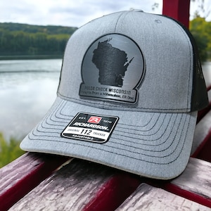 May include: A gray and black trucker hat with a silver patch featuring the outline of the state of Wisconsin. The patch has the text "PULSE CHECK WISCONSIN" and "noughts from a Milwaukee, ER Doc". The hat is adjustable and has a Richardson 112 label.